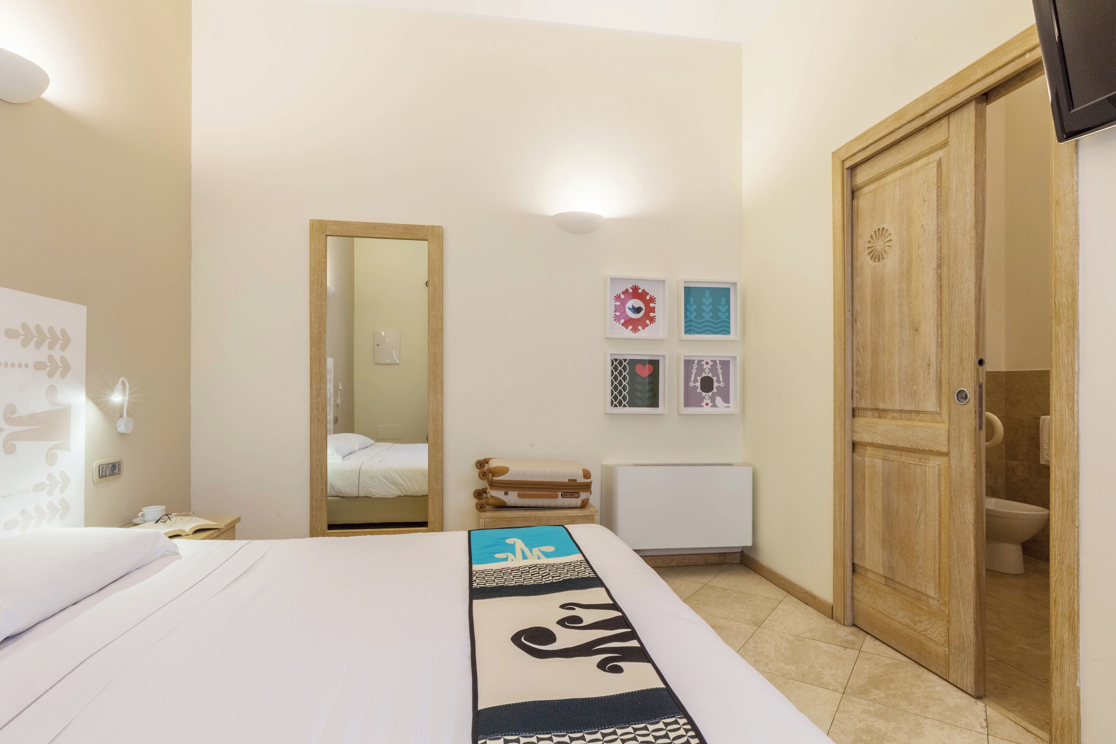 Ein Hotelzimmer mit einem Bett, einem Holzschrank, einem Spiegel, einem kleinen Tisch mit einem Buch und einer Wand mit gerahmten Bildern.
