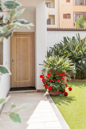 A wooden door, a potted plant with red flowers, and a well-maintained lawn. - Hotel Domomea (Alghero)