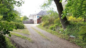 A gravel driveway leading to a white house with a blue sign, surrounded by lush greenery and trees. - Relais des Caillères (Saint-Pavace)
