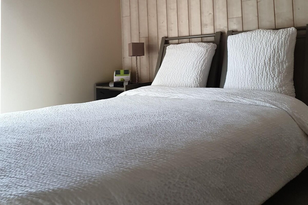 Camera da letto con soffitto in legno, un letto con biancheria bianca, un comodino con una lampada e una trave di legno sulla parete.