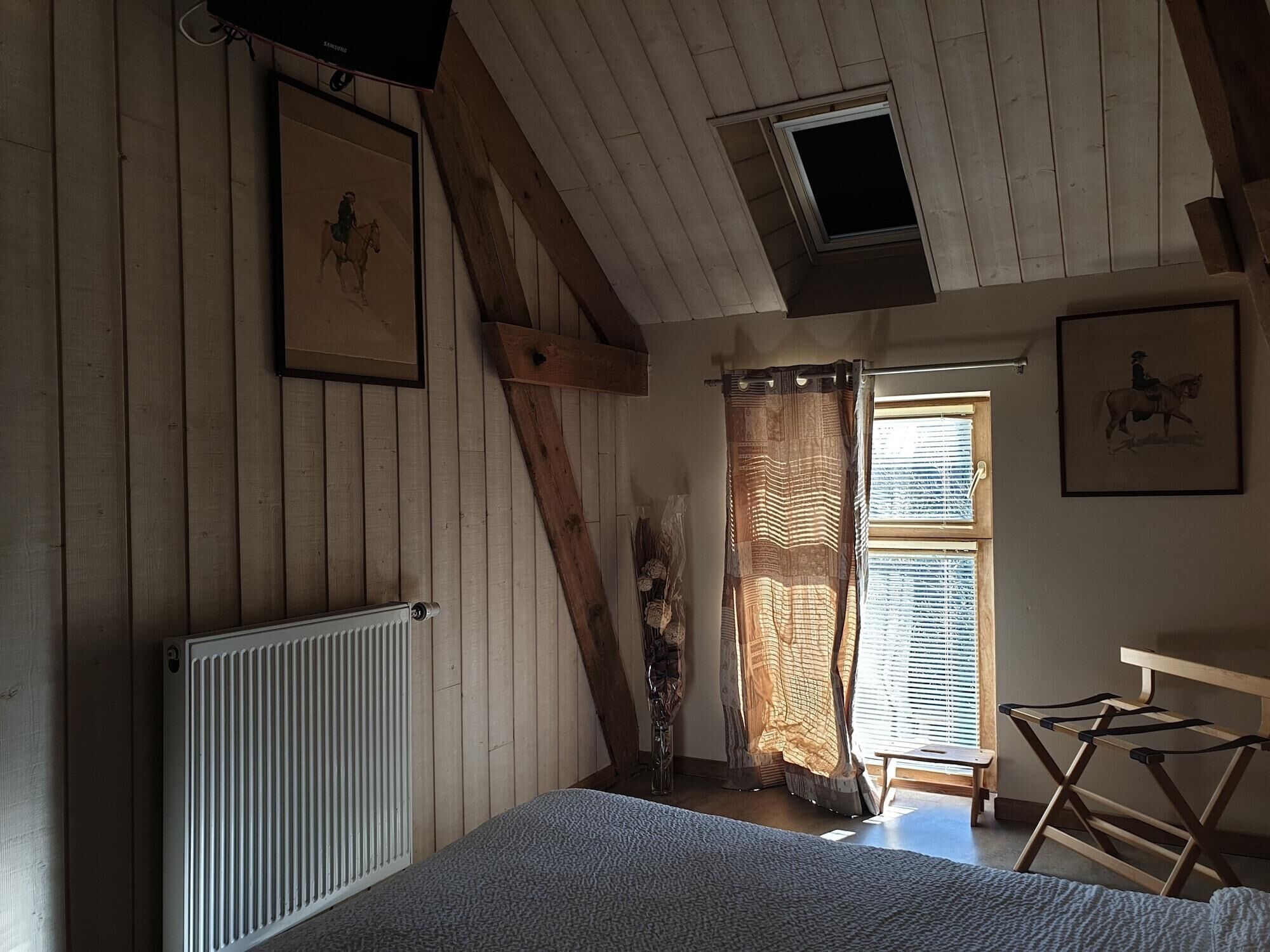 Une pièce aux murs en bois, avec un plafond en pente, un radiateur, un lit, une fenêtre avec des stores et une petite table.