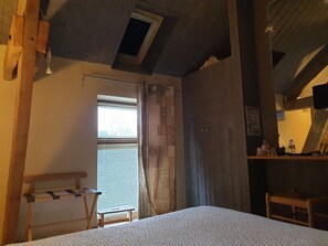 Una habitación con cama, un banco de madera, una mesita, una ventana con persianas y un techo de madera con tragaluz.