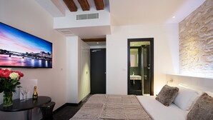 Una habitación de hotel moderna con una cama grande, un televisor de pantalla plana, una mesita con un jarrón de flores y vistas a la ciudad.