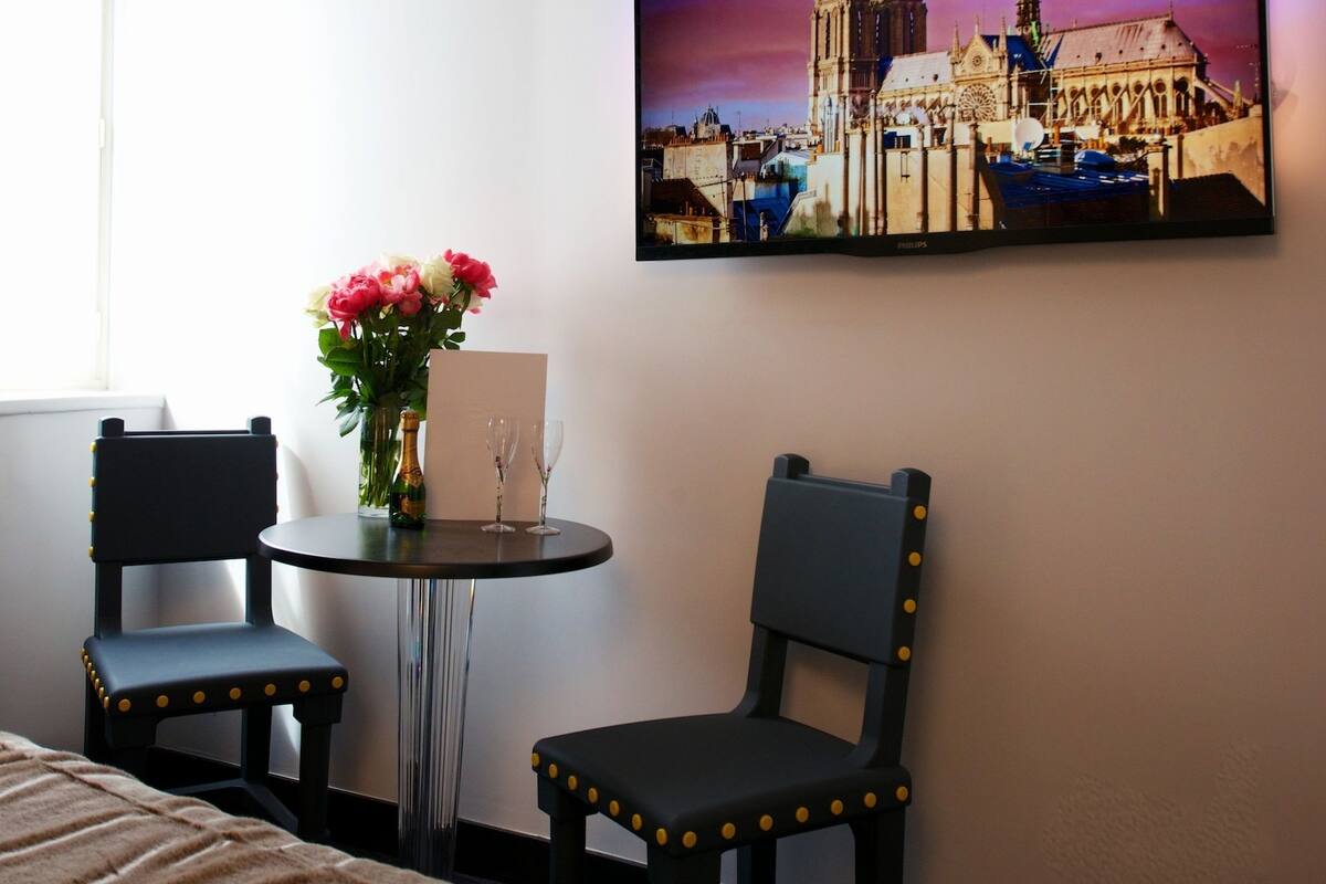 Habitación de hotel con cama, una pequeña mesa con sillas, un jarrón de flores y un cuadro con una vista urbana en la pared.