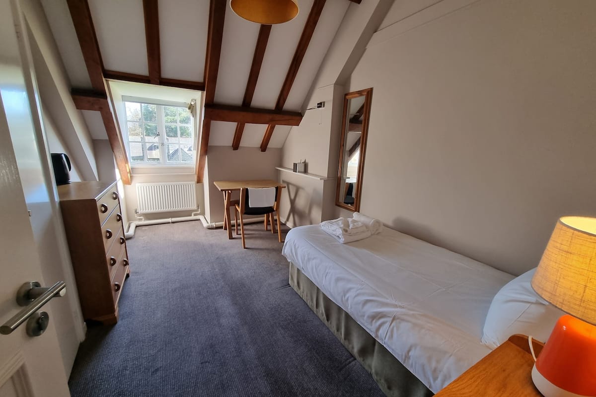 A compact room with a bed, a desk, and a chair. There is a window providing natural light and a view of the outside.