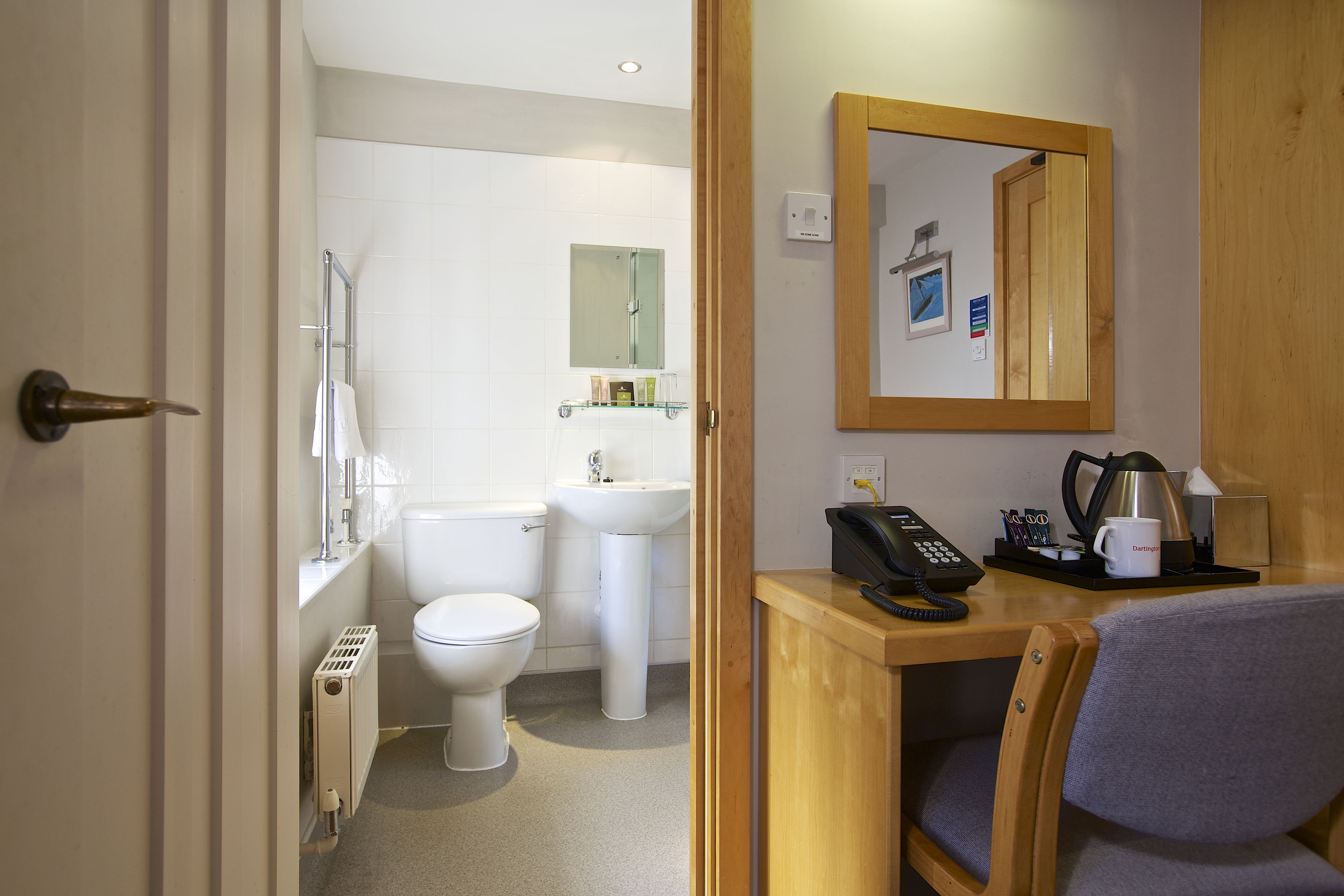 A bathroom with a toilet, sink, and shower. There is a wooden desk with a chair, a telephone, and a coffee maker.