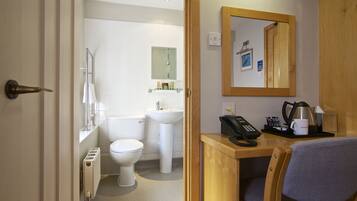 A bathroom with a toilet, sink, and shower. There is a wooden desk with a chair, a telephone, and a coffee maker.