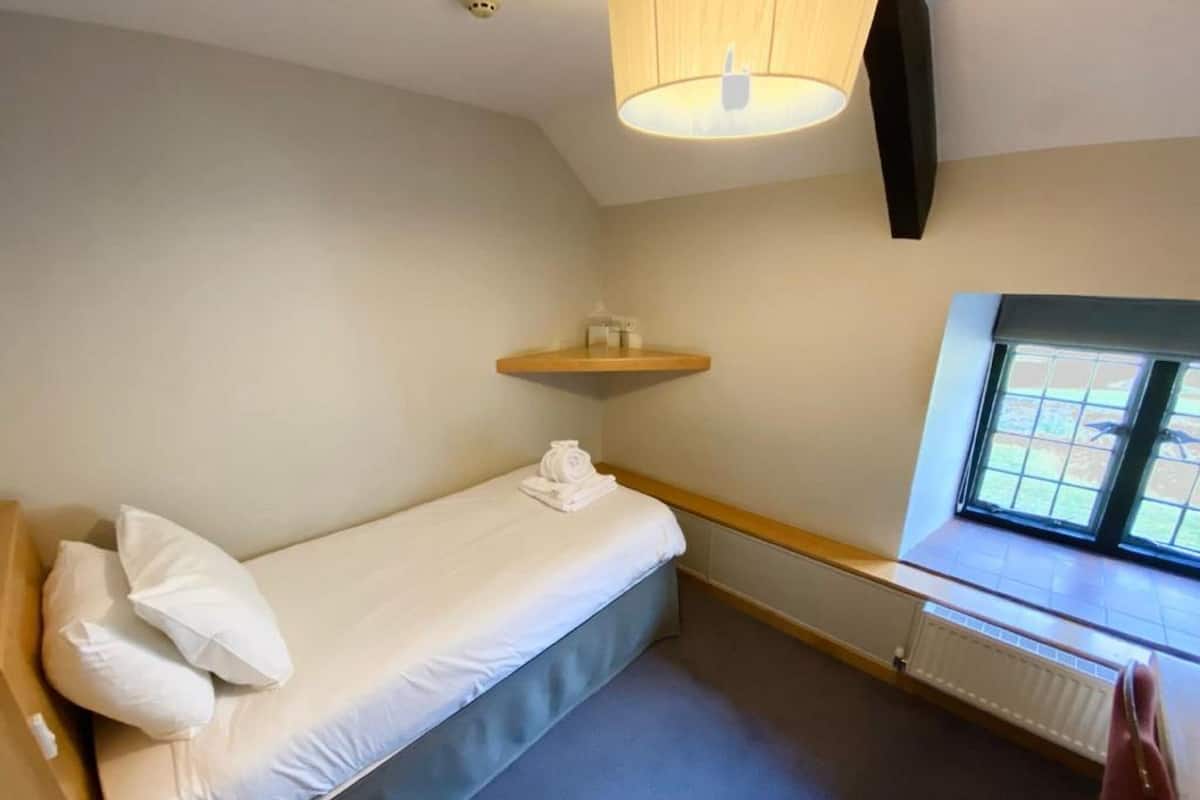 A small, single-bed room with a wooden headboard, a bedside table with a lamp, a window with a view, and a radiator.