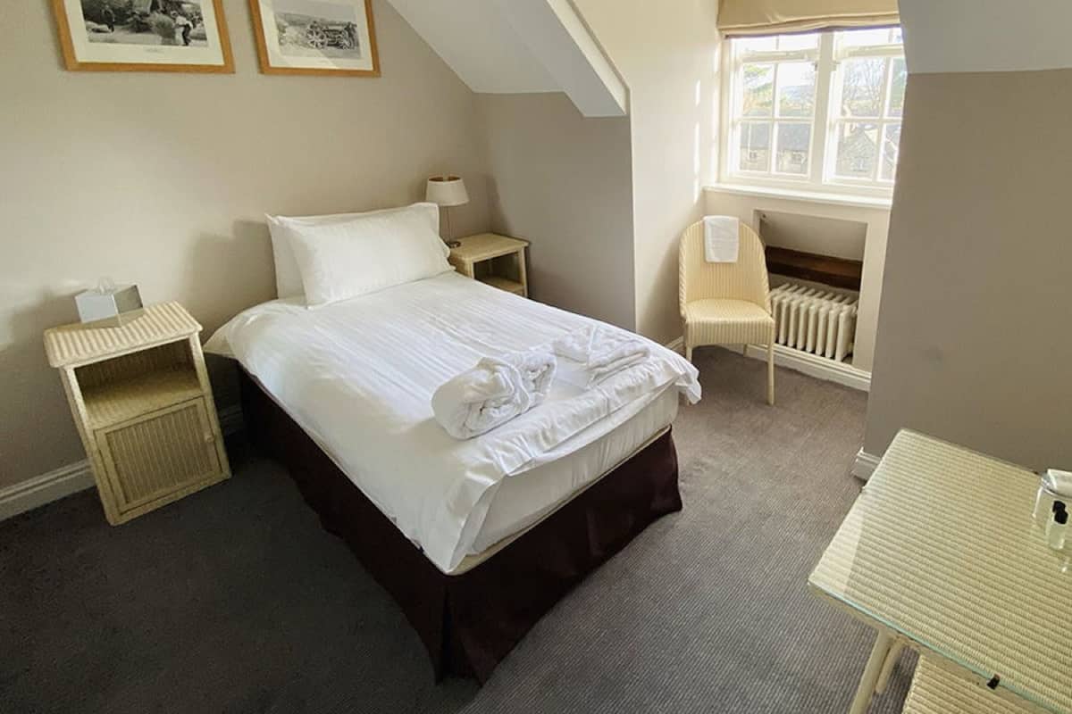 A neatly made bed with white linens and towels, a bedside table with a lamp, a chair, and a radiator in a room with a sloped ceiling and a window.