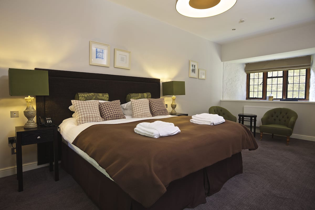 A neatly made bed with a brown bedspread and pillows, a bedside table with a lamp, a chair, and a window with blinds.