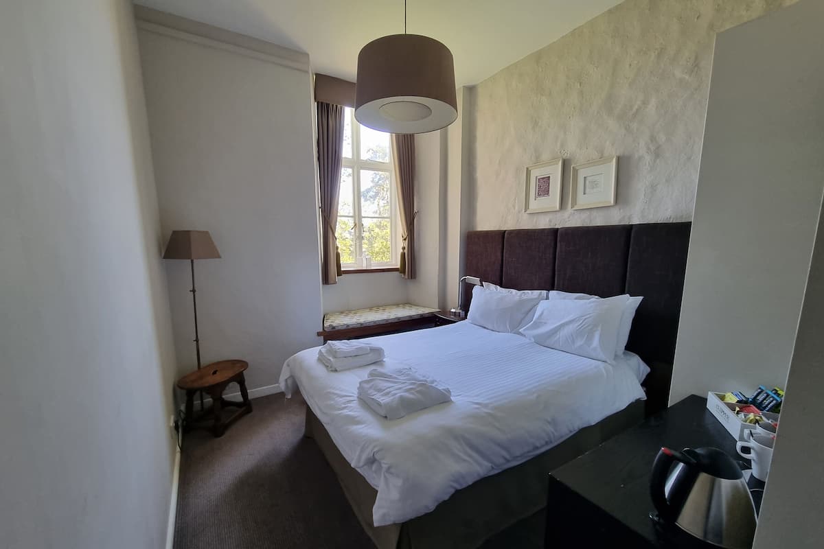 A hotel room with a large bed, a bedside table with a kettle, a small stool, a floor lamp, and a window with curtains.