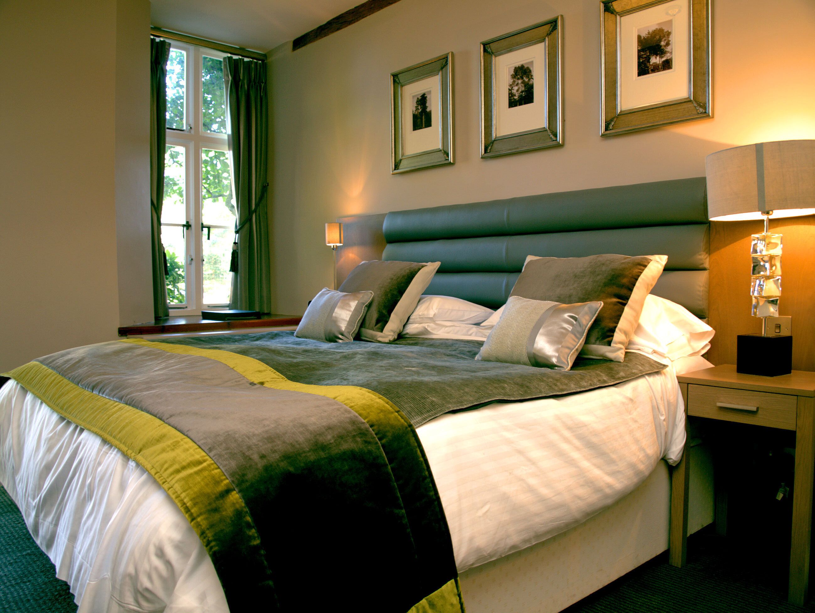 Una cama bien hecha con edredón gris y blanco, una manta de color verde y amarillo, y varias almohadas. Hay cuadros enmarcados en la pared sobre la cama, y una lámpara de noche en la mesita.