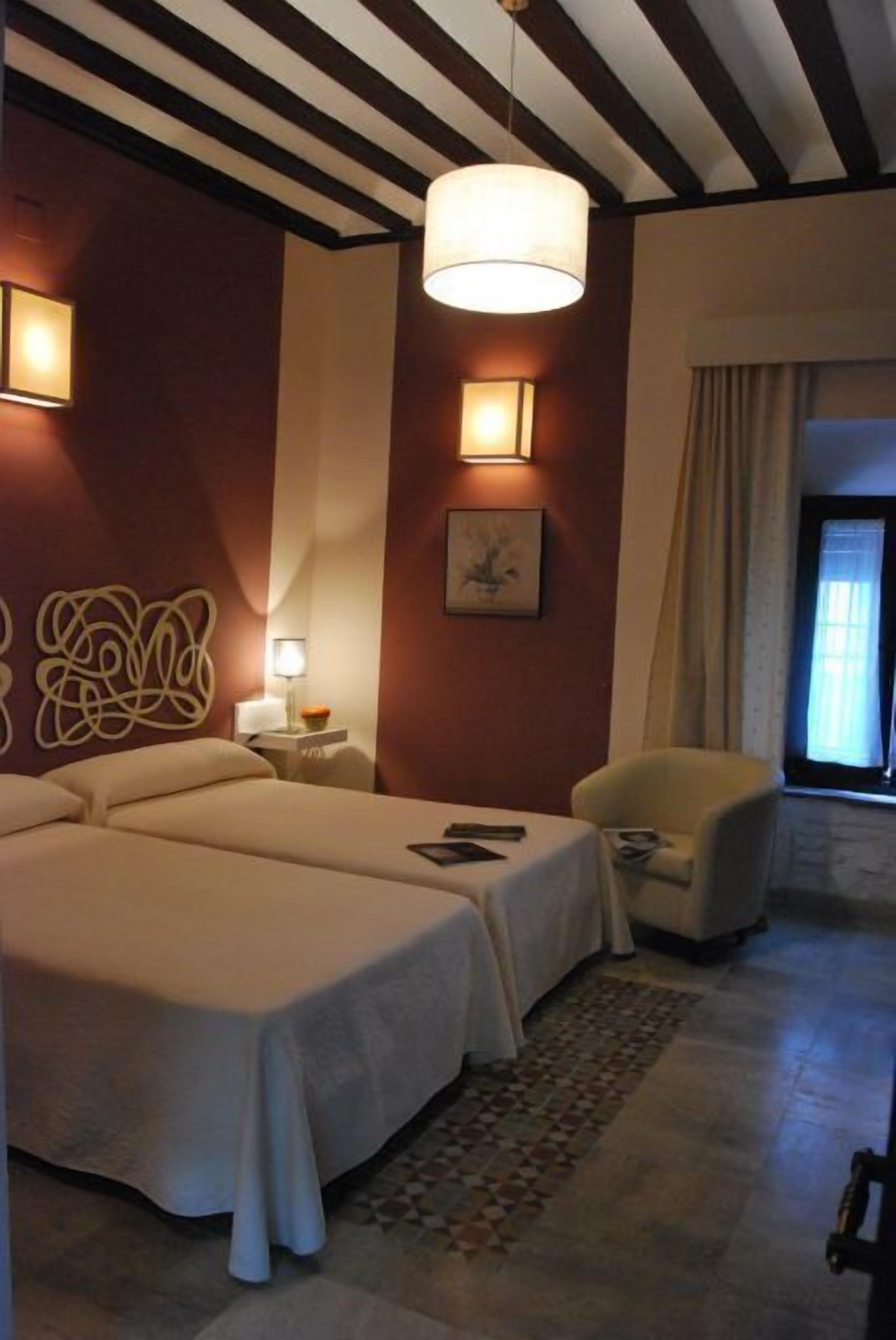 A hotel room with two beds, a sitting area with a chair, a lamp, and a painting on the wall.