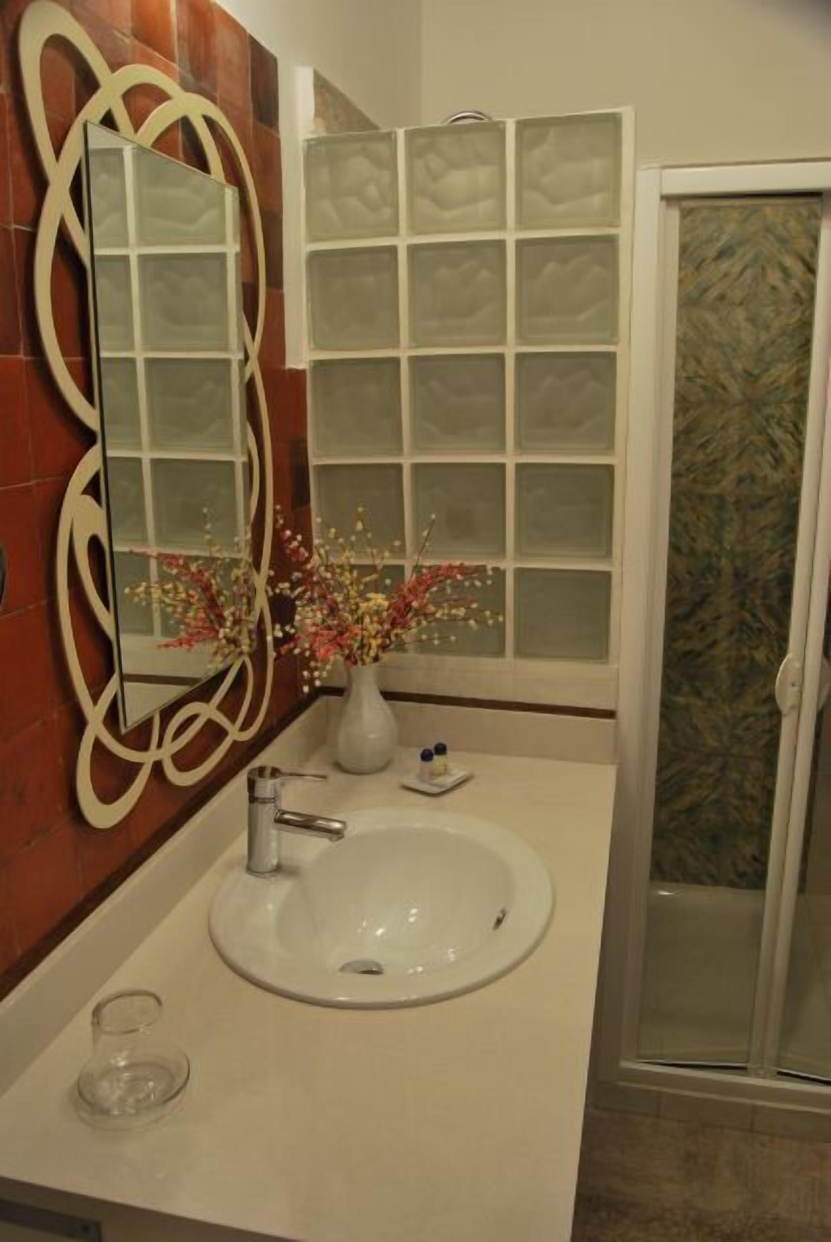 A bathroom with a white sink, a mirror with a decorative frame, a vase with flowers, and a frosted glass window.