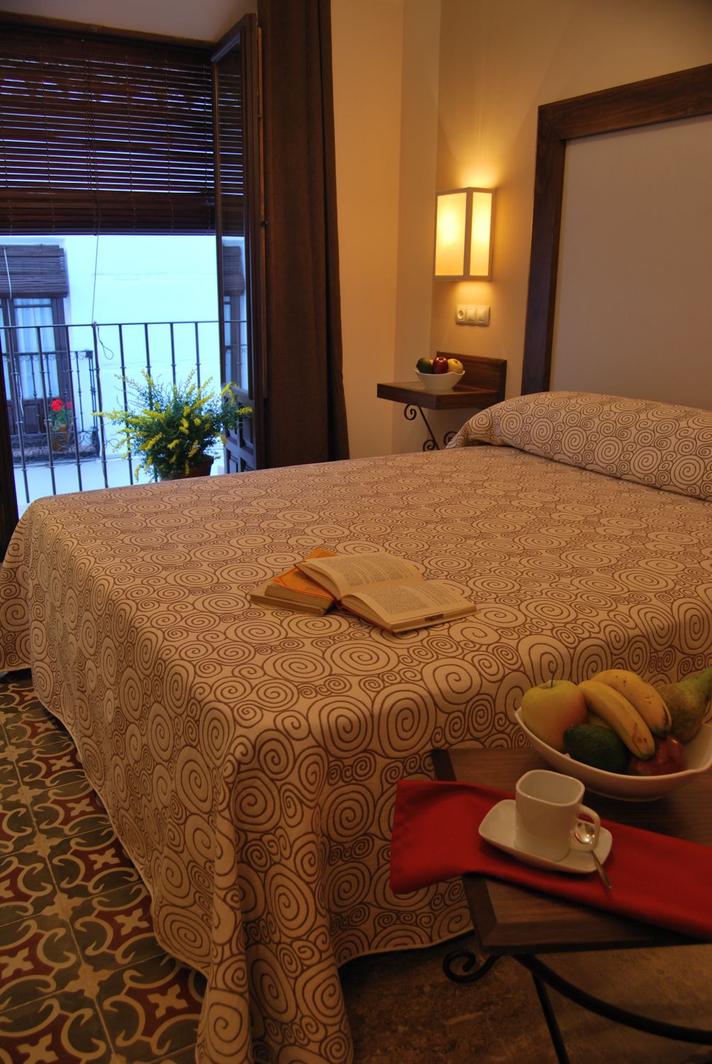 Habitación de hotel con cama, mesita de noche con lámpara, frutero y ventana con persianas.