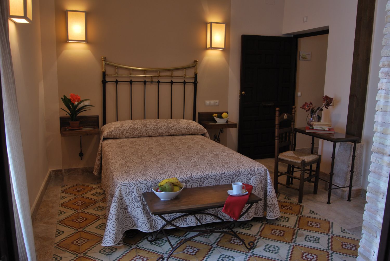 Un dormitorio ordenado con una cama, mesita de noche y una pequeña mesa con un frutero y una taza.
