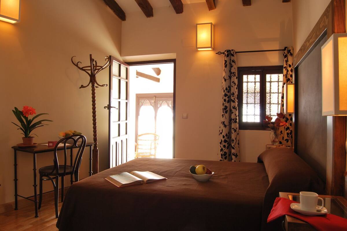A bedroom with a bed, a bedside table with a cup, a chair, a desk with a plant, and a window with curtains.