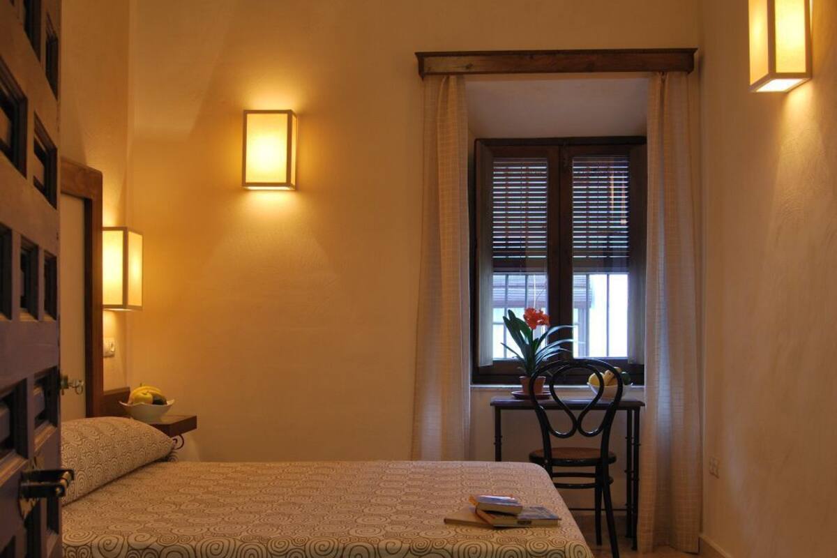 A hotel room with a bed, a desk, a chair, a window with blinds, and a potted plant.
