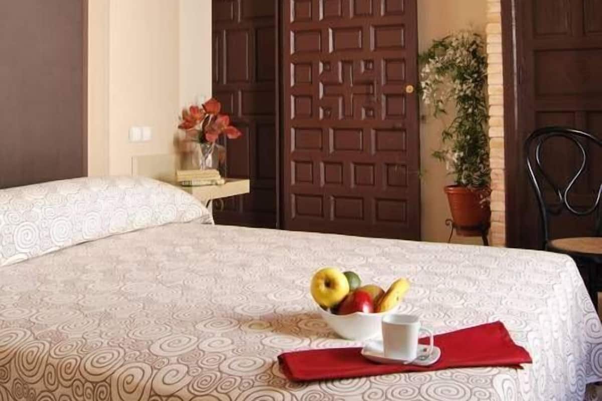 A hotel room with a bed, a bedside table with a vase of flowers, a fruit bowl, and a red napkin.