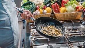 A kitchen with a person cooking noodles on a stove, surrounded by baskets of fresh vegetables. - Hotel La Fenice (Castel di Sangro)