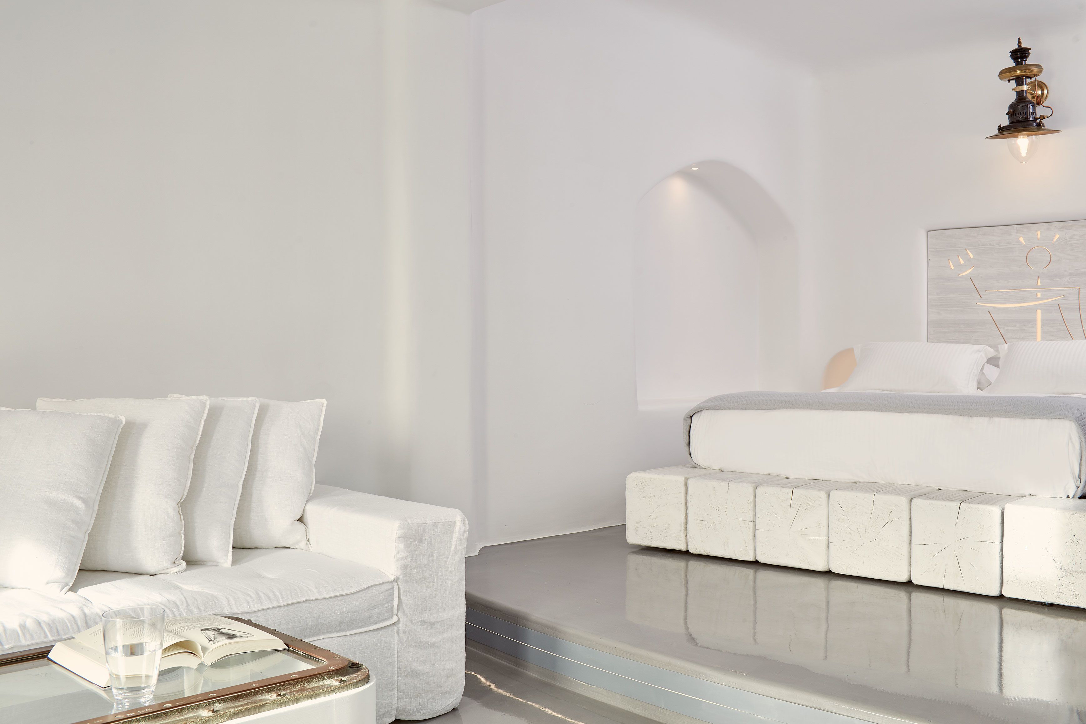 Quarto moderno e minimalista, com um sofá branco, uma mesa de cabeceira com um livro e um copo, e uma cama com cabeceira branca.