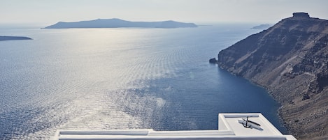 A rooftop terrace with sun loungers overlooking a calm sea and a mountainous landscape.