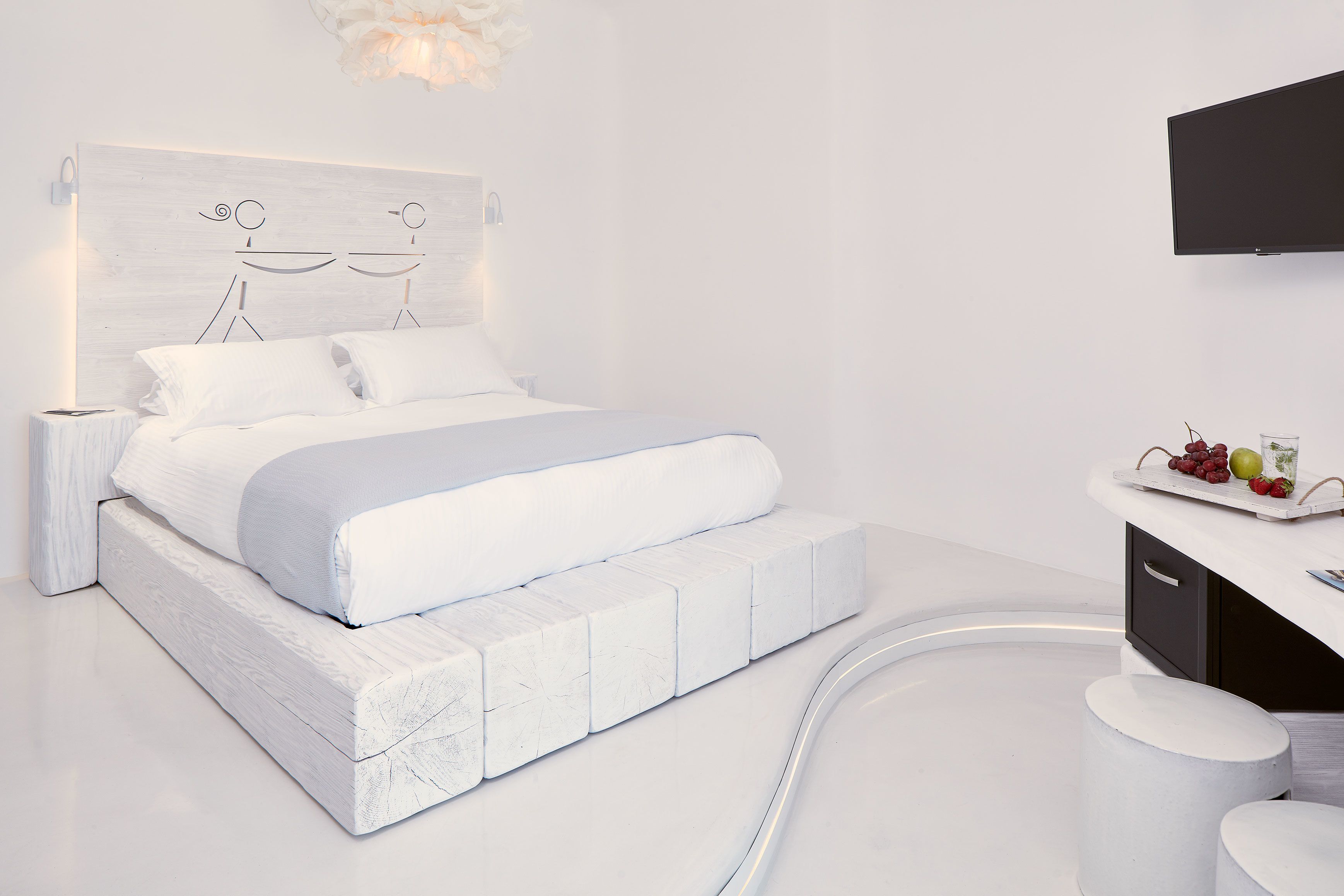 Un dormitorio moderno con cama blanca, cabecero de diseño minimalista, mesita de noche y televisor de pantalla plana montado en la pared.