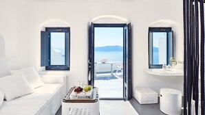 A modern, minimalist room with a white sofa, a small table with fruits, a view of the sea, and a white bench. - Homeric Poems (Santorini)