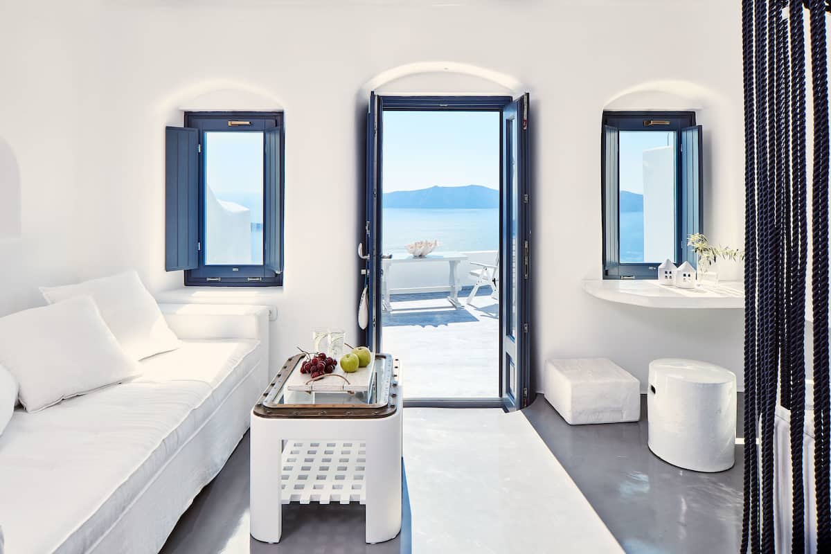 A modern, minimalist room with a white sofa, a small table with fruits, a view of the sea, and a white bench.
