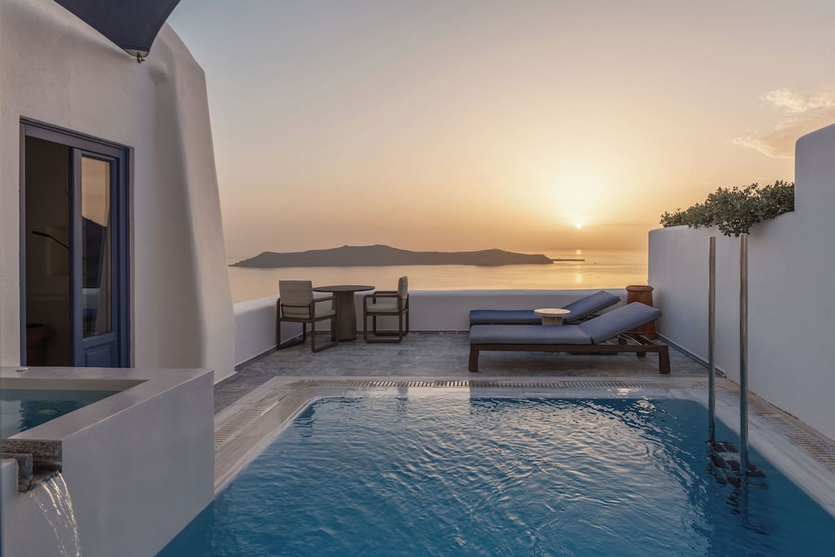 A rooftop pool area with lounge chairs and a table, overlooking a sunset over the sea.