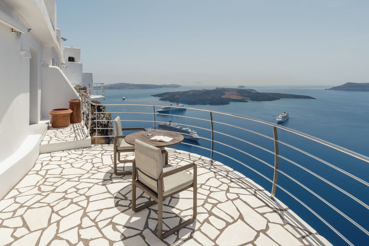 A balcony with a stone floor, two chairs, a small table, and a railing overlooking a body of water with boats and distant islands.