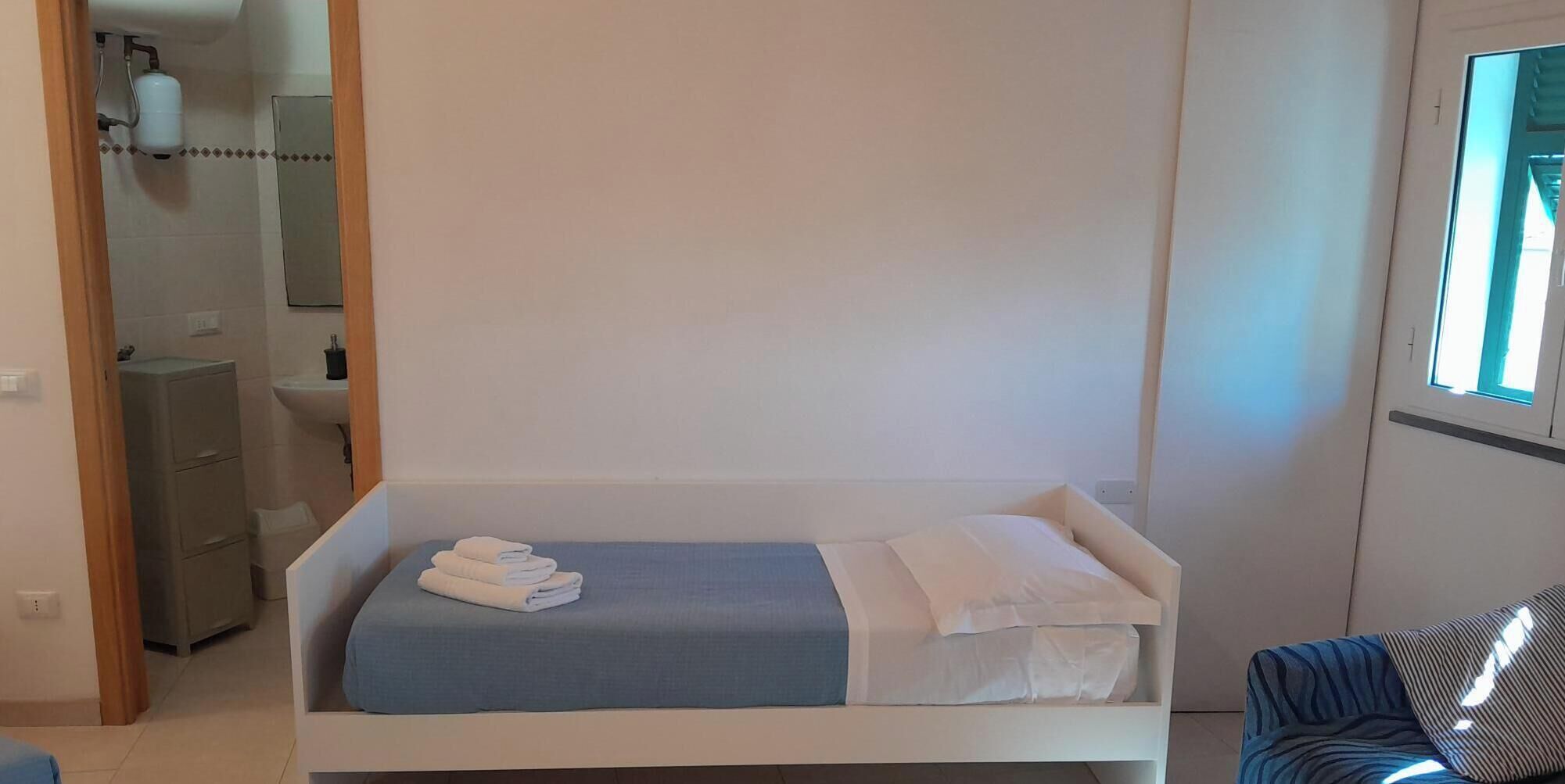 A single bed with a blue bedspread and white pillows, a small bedside table, and a window with a view.