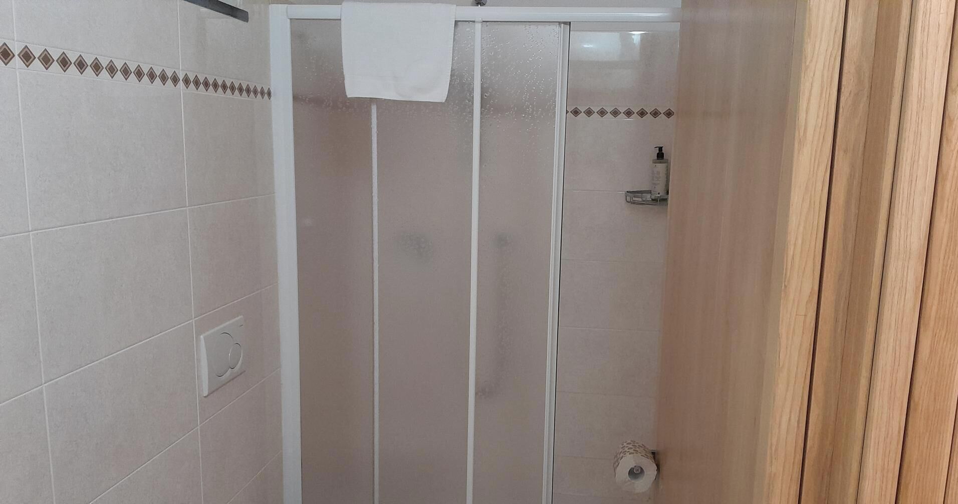 A shower with a white towel hanging on the door, a soap dispenser on the wall, and a toilet paper holder.