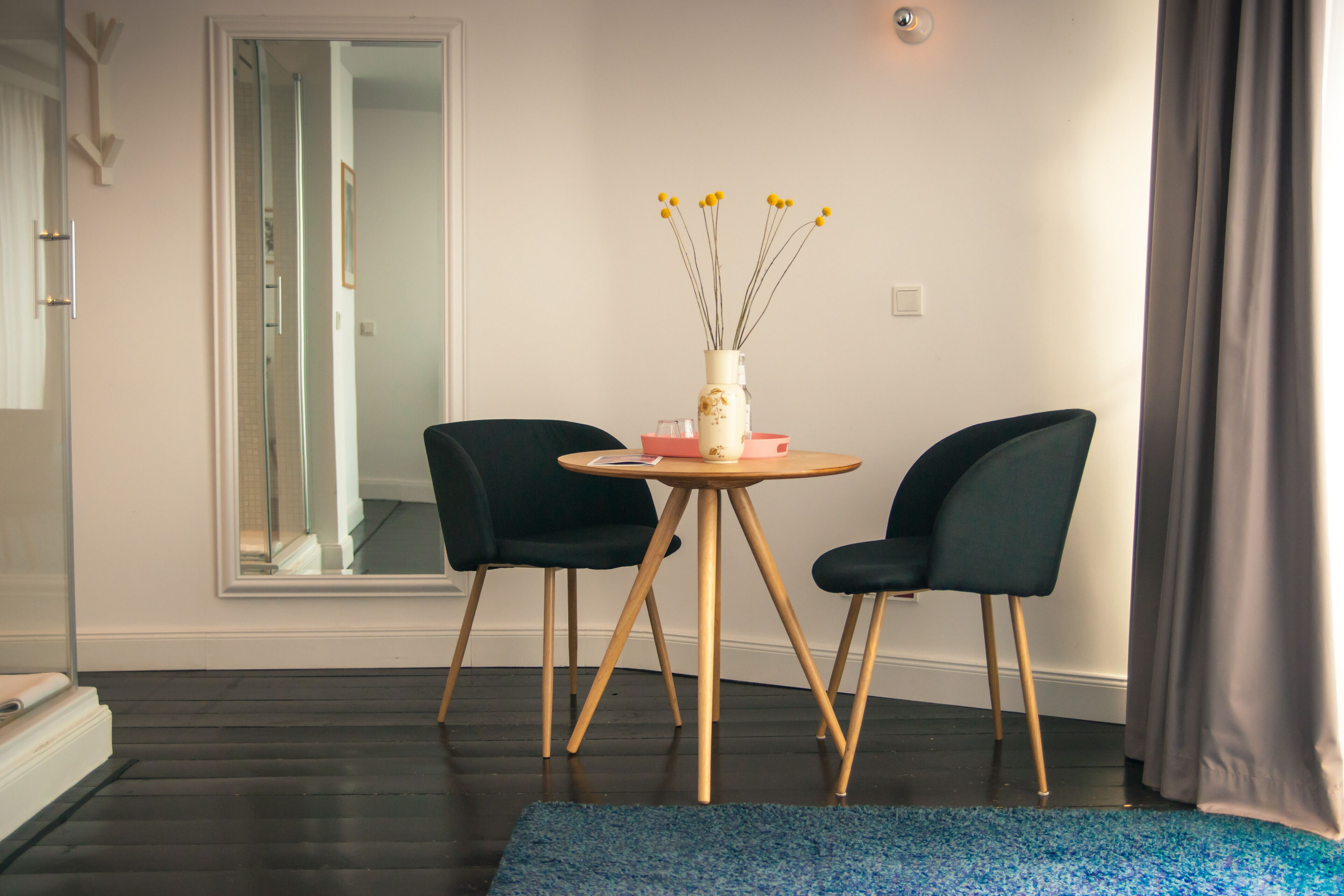 Ein modernes Zimmer mit dunklem Holzboden, einem runden Tisch mit zwei Stühlen, einer Vase mit gelben Blumen und einem großen Spiegel.