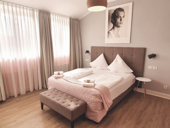 Un lit bien fait, recouvert d’une couverture rose, une table de chevet et un banc dans une chambre d’hôtel moderne dotée d’une grande fenêtre et de rideaux.