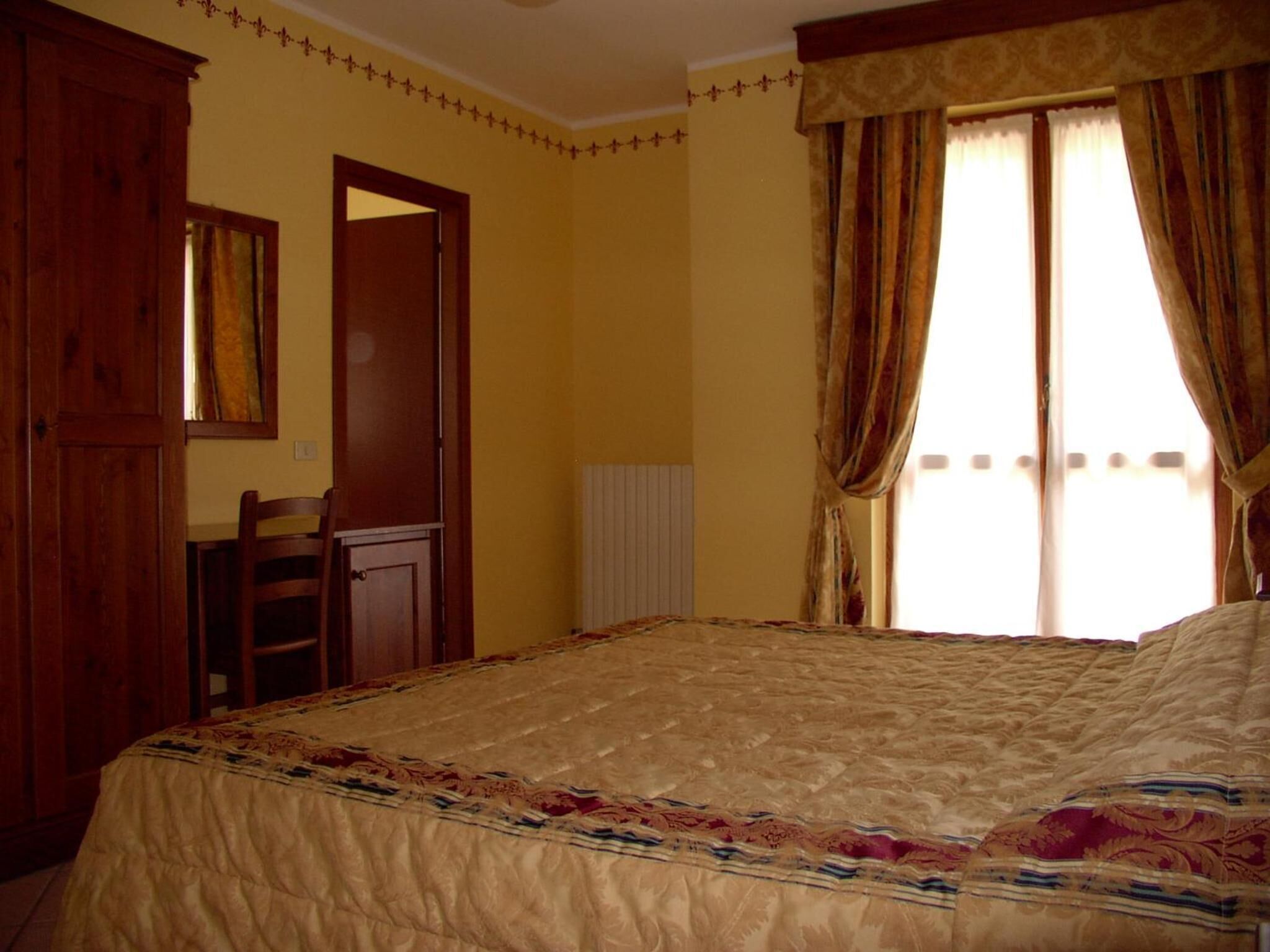 A bedroom with a large bed, a wooden wardrobe, a desk, and a window with curtains.