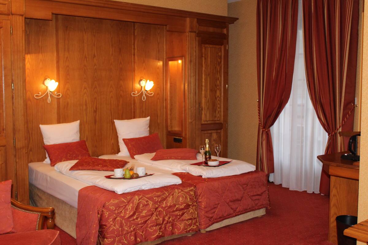 Ein Hotelzimmer mit zwei Betten, roter Bettwäsche, einem hölzernen Kopfteil, einem Nachttisch mit Flasche und Gläsern sowie einem roten Vorhang.