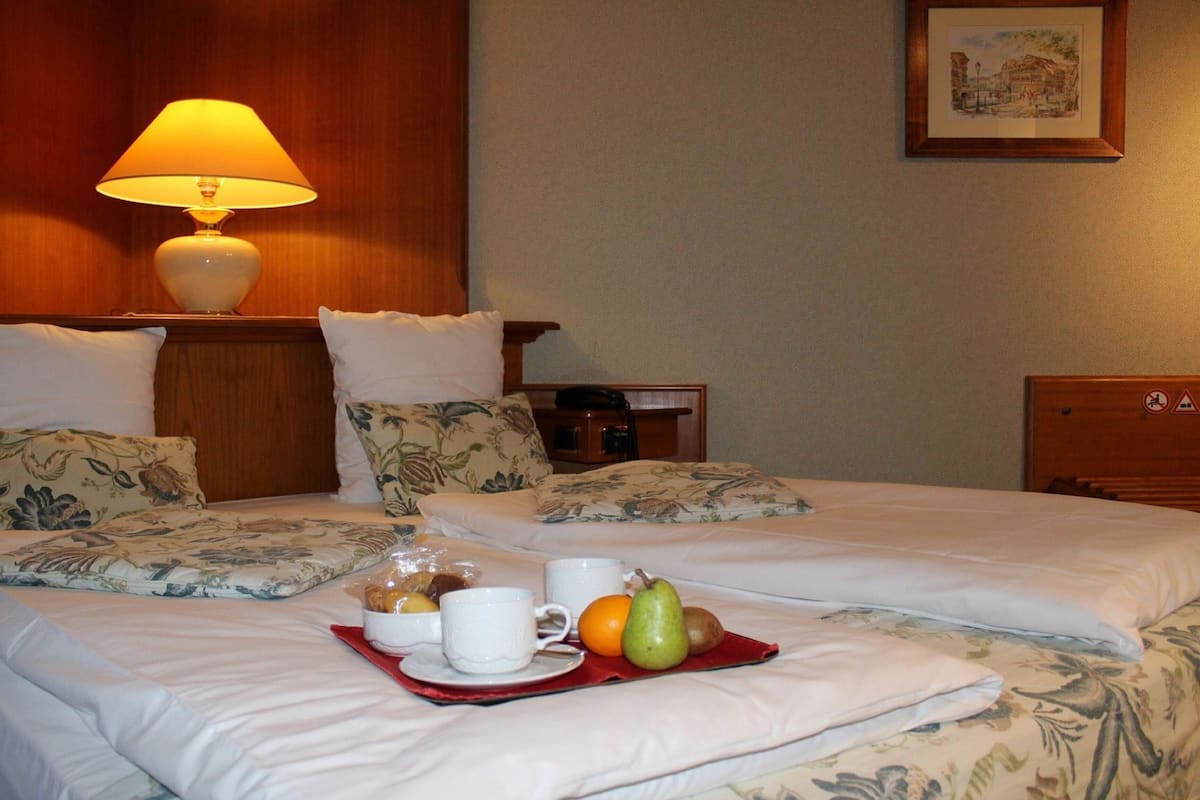 Ein Hotelzimmer mit zwei Betten, einem Holz-Kopfteil, einer Lampe und einem Tablett mit Früchten und Tassen.