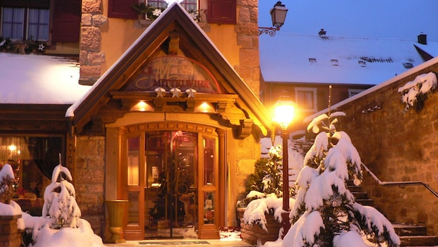 Un bâtiment recouvert de neige, avec une entrée en bois, des marches menant à la porte et des arbres également couverts de neige.