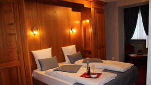 Une chambre d’hôtel avec un grand lit, des boiseries, deux lampes fixées au mur et une petite table sur laquelle se trouvent une bouteille et un verre.