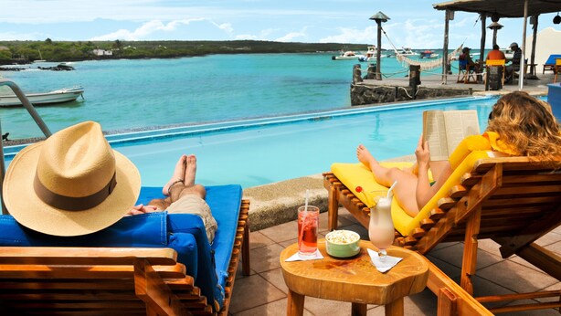 A poolside area with lounge chairs, a hat, a drink, and a view of the sea and boats. - Hotel Solymar (Puerto Ayora)