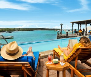 A poolside area with lounge chairs, a hat, a drink, and a view of the sea and boats. - Hotel Solymar (Puerto Ayora)
