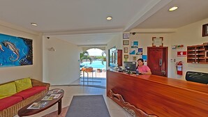 A reception area with a wooden reception desk, a receptionist, a colorful painting on the wall, a wicker sofa, and a view of the pool through the glass door. - Hotel Solymar (Puerto Ayora)