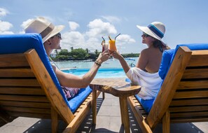 Dos personas sentadas en sillones de madera con cojines azules, sosteniendo bebidas, y contemplando una piscina con vistas despejadas al océano y follaje en el fondo.
