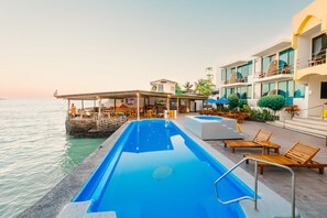Zona junto a la piscina con sillones, un bar y un edificio con balcones con vistas al agua.