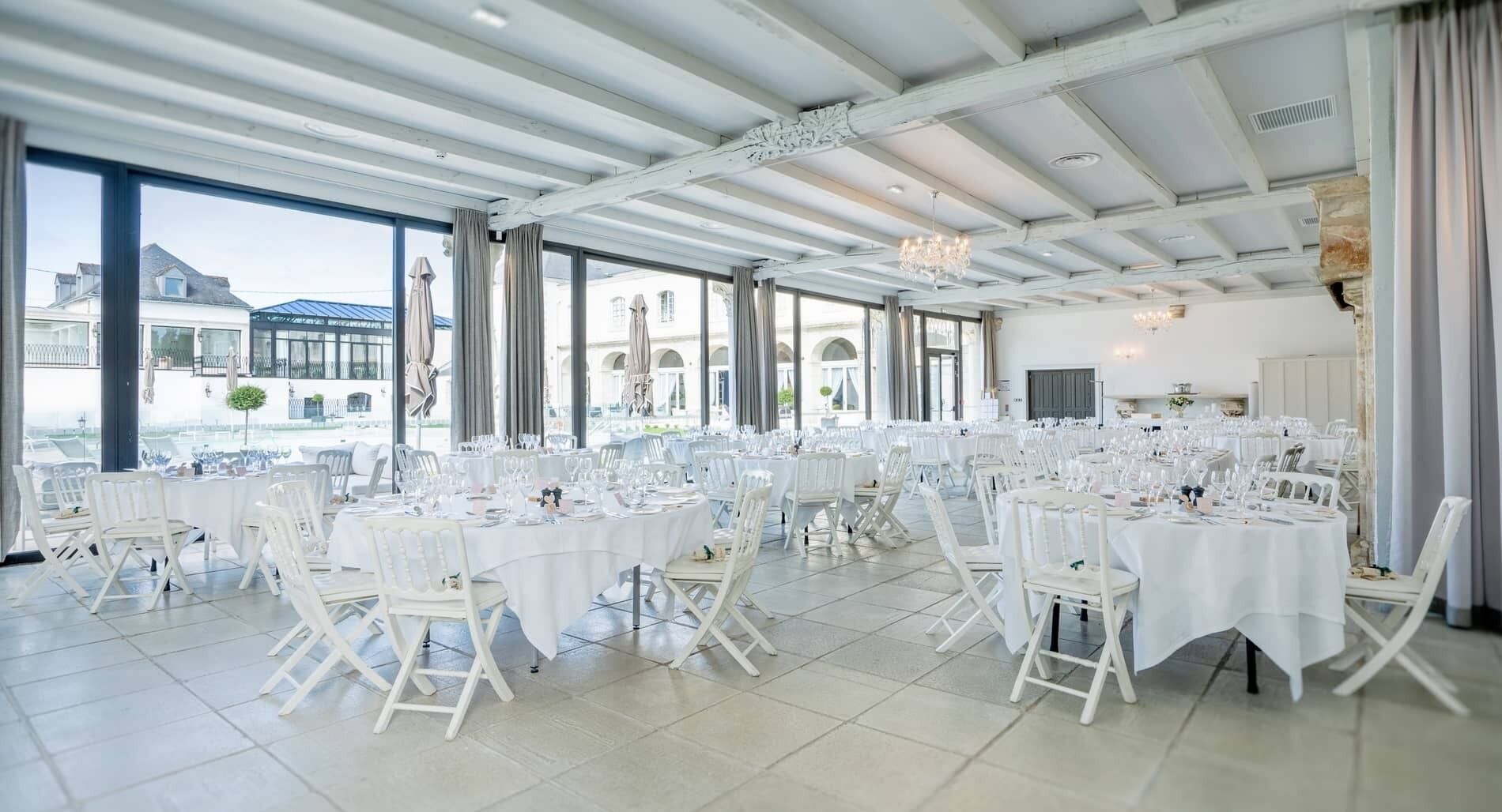 A spacious indoor venue with multiple round tables set for dining, large windows offering views of the outside, and a high ceiling with exposed beams.