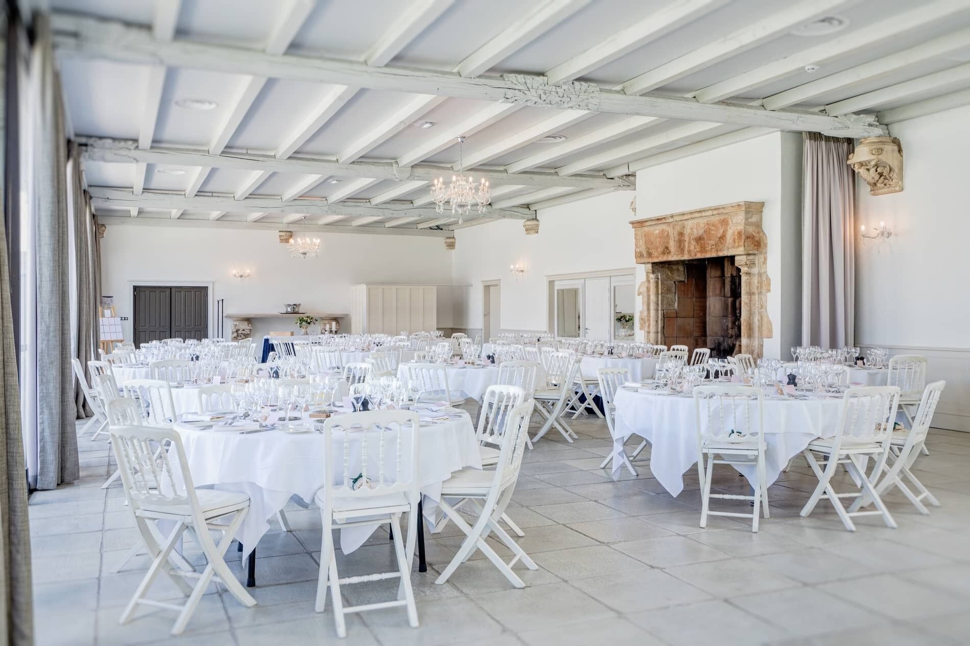 Um amplo salão de banquetes com mesas e cadeiras brancas, lustres e uma lareira de pedra.