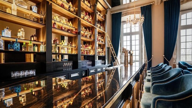 Una zona de bar con estantes llenos de botellas de licor, una barra pulida y filas de sillas tapizadas en azul.