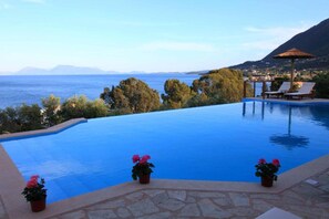 A swimming pool with a view of the sea and mountains, lounge chairs, and potted plants.