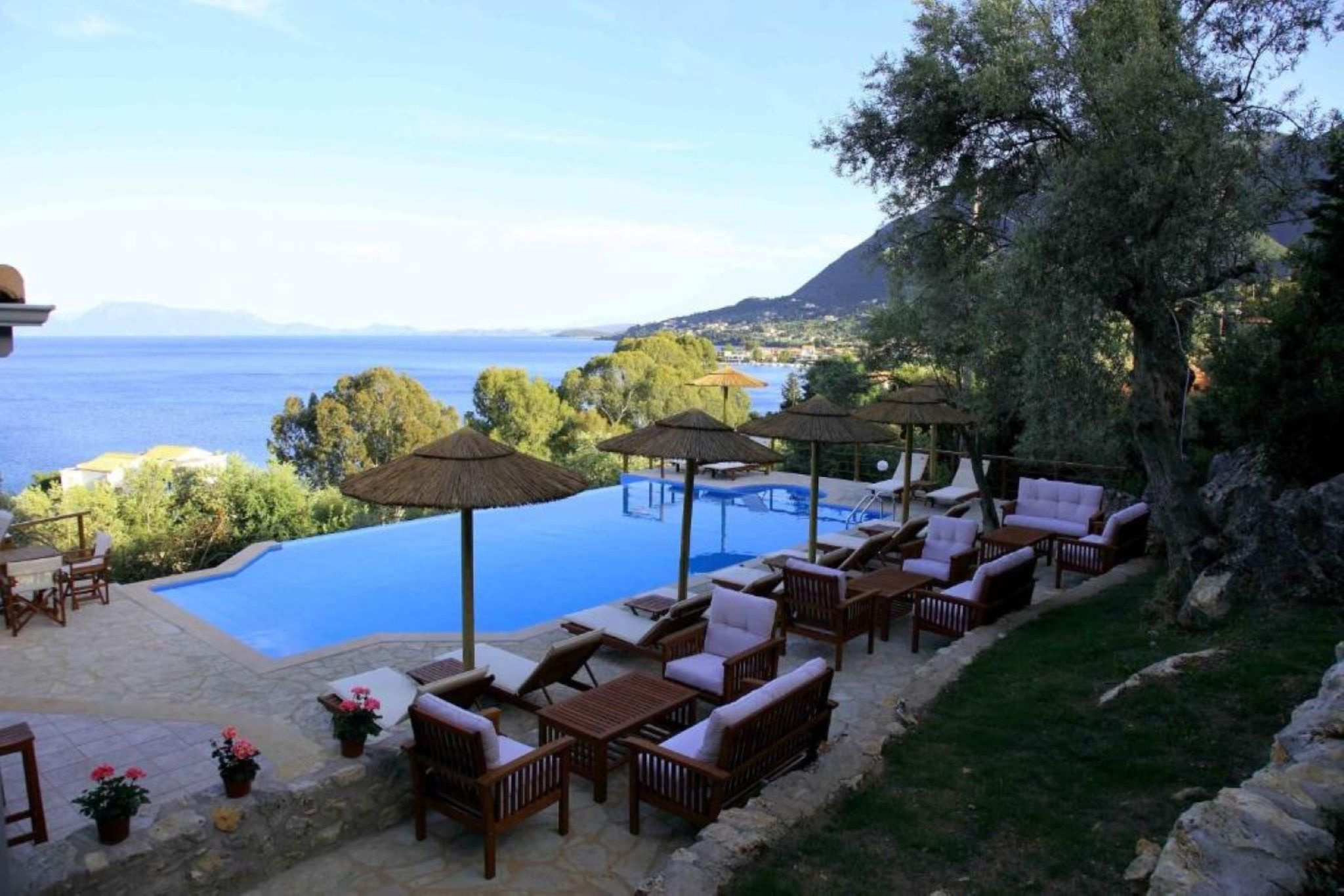 Zona de piscina exterior con sillones de descanso y sombrillas, con vistas a un cuerpo de agua y un paisaje montañoso.